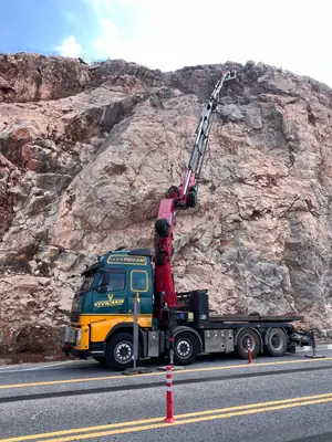 Γερανοφόρο όχημα με εκτεταμένο βραχίονα σε εργασία σε βραχώδες πρανές δίπλα σε δρόμο